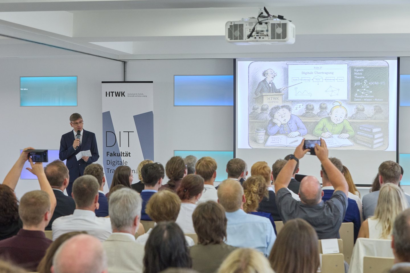 Redner mit Mikro und Präsentation vor vollbesetzten Stuhlreihen