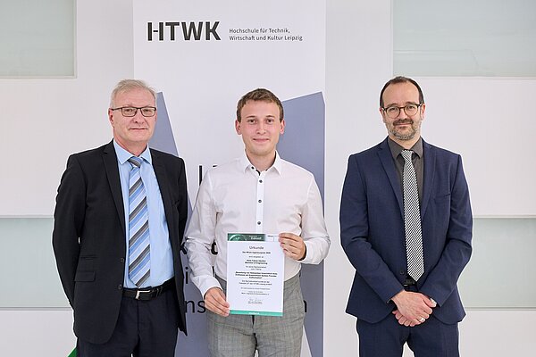 Bachelor (Eng.) Niels Fabien Häußler erhielt an der HTWK Leipzig den IfKom-Preis 2025 drei Personen vor Plakat, davon einer mit Urkunde