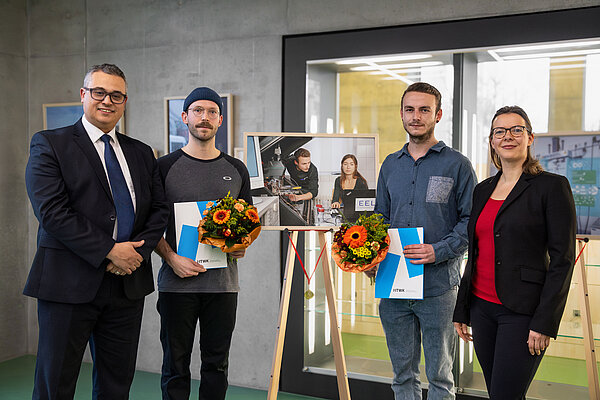 Gewinner Forschungsperspektiven 2024 drei Männer und eine Frau stehen vor einem Bild auf einer Staffelei, zwei halten Urkunden und Blumen in den Händen