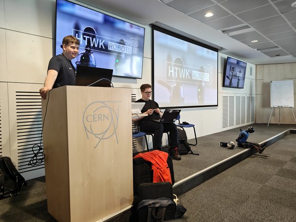 Vortrag der HTWK Robots im CERN (v.l.n.r. David Schulte, Max Polter, Ganymed) (c) HTWK Robots Person steht beim Vortrag am Pult vor einer Wand mit drei Monitoren. Daneben sitzt eine weitere Person mit Laptop auf den Knien und ein humanoider Roboter.