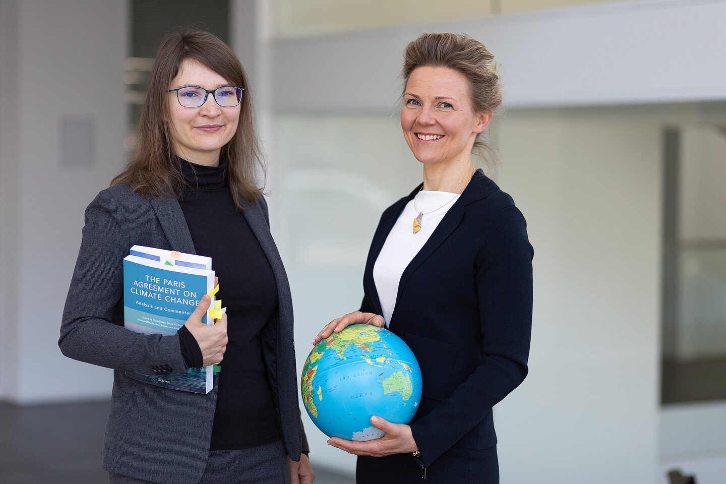 Zwei Frauen mit Büchern und einem Globus in der Händen lächeln in die Kamera.