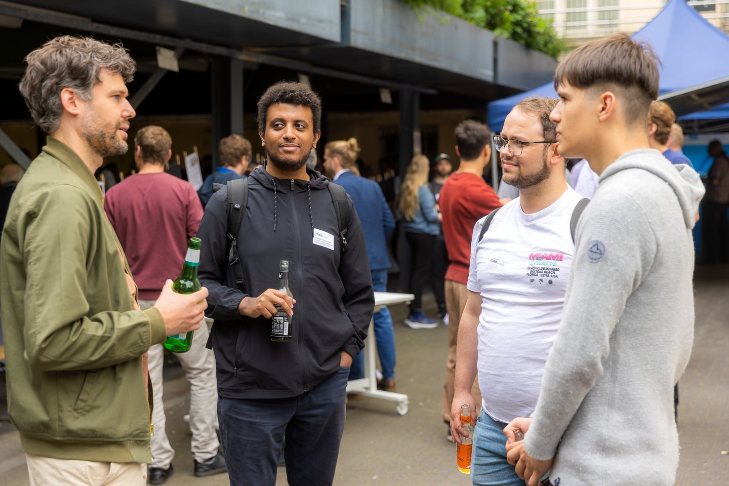 Vier Männer stehen nebeneinander, unterhalten sich und halten jeweils ein Getränk in der Hand. Im Hintergrund sind weitere Gruppen von Menschen zu sehen